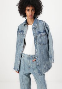 Denim jakna v svetlo modri barvi z ohlapnim krojem, velikimi žepi na prsih, pritisnimi gumbi in rahlo razfrčanim robom, nosena čez belo majico.