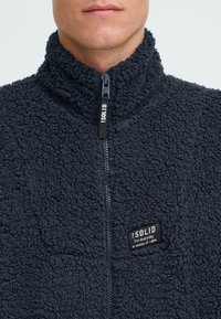 Fleece zip-up jakke i mørkegrått med høy krage. Har en strukturert overflate og en liten svart etikett på brystet.