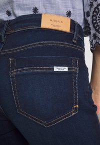 Mörkblå denimjeans med en synlig läderetikett i midjan. Har kontrasterande gul söm och en bakficketikett.