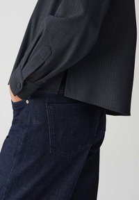 Persona che indossa una maglietta nera a maniche lunghe con maniche a doppio risvolto e jeans in denim blu scuro, con una mano nella tasca anteriore.