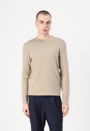Beige langärmliger Pullover mit Rundhalsausschnitt. Weicher, strukturierten Stoff. Kombiniert mit navyblauen Hosen, die eine lockere Passform und Falten haben.