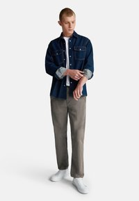 Camicia di denim blu scuro con polsini azzurri, indossata sopra una maglietta bianca. Pantaloni beige a gamba dritta e sneakers bianche completano il look.