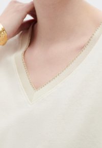 Top w serek w jasnym kremie, z teksturowanym złotym szwem wzdłuż dekoltu. Miękka tkanina o gładkim wykończeniu i luźnym kroju.