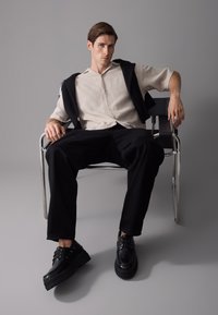 Zapatos de plataforma negros, pantalones negros y una camiseta de manga corta beige con rayas verticales, llevada con un suéter negro colocado sobre los hombros.