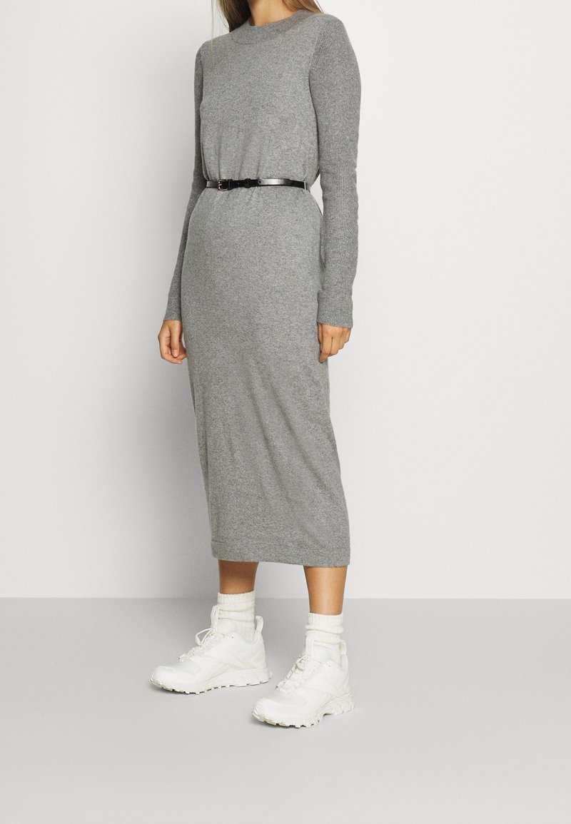 Femme portant une robe midi en maille grise à manches longues avec une ceinture noire, des chaussettes blanches hautes et des baskets blanches épaisses, debout contre un mur uni.