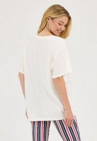 Balts oversized t-krekls ar īsām piedurknēm, gluda tekstūra un apaļa kakla izgriezuma. Valkāts kopā ar daudzkrāsu svītrainām, pieguļošām biksēm.