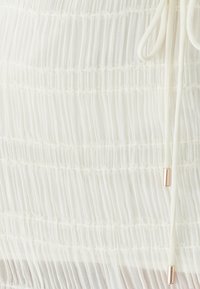 Weißes, plissiertes Stofftop mit zartem Spitzenbesatz, ausgestattet mit einem Tunnelzugverschluss und metallischen Akzenten zur Anpassung. Insgesamt weiche Textur.