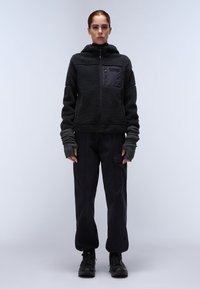 Chaqueta con capucha forrada de forro polar negra y bolsillo con cremallera en el pecho; combinada con pantalones negros sueltos y guantes sin dedos texturizados. Calzado negro.