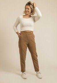 Pull en tricot blanc à manches courtes associé à un pantalon ajusté marron avec un accent de bouton et des baskets blanches. Fond clair.