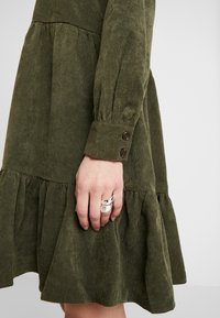 Robe en velours côtelé vert avec une jupe à volants, des manches longues, des poignets boutonnés et une finition texturée. Main portant des bagues en argent visible.