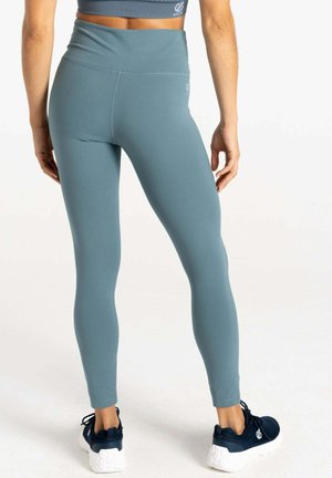 Femme portant un legging bleu taille haute et des chaussures de sport noires, debout de dos à la caméra sur un fond blanc.