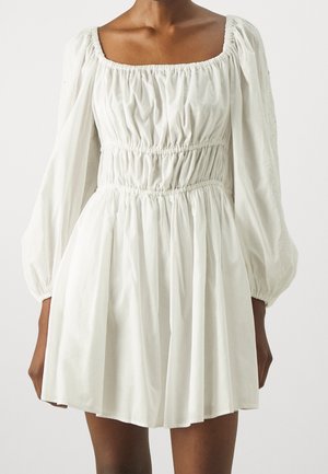 Mujer que lleva un vestido blanco con mangas largas abullonadas, escote cuadrado y cintura y falda fruncidas.
