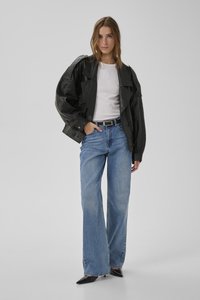 Vrouw draagt een oversized zwarte leren jas, wit shirt, wijde blauwe jeans en zwarte puntige hakken, staand met één hand in haar zak.