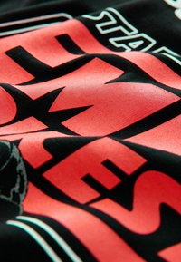 Tricou din material textil negru, cu litere îndrăznețe de culoare roz și detalii grafice, având o texturăfină și accente albe parțial vizibile.