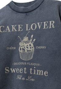 Sudadera gris con un gráfico de cupcake adornado con cerezas y el texto "AMANTE DEL PASTEL", "Sabor Delicioso" y "Tiempo Dulce" en bordado metálico.