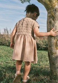 Světle růžové bavlněné šaty s háčkovaným vrškem, krátkými rukávy, texturovanou sukní a zapínáním na knoflík vzadu; dítě stojící u stromu.
