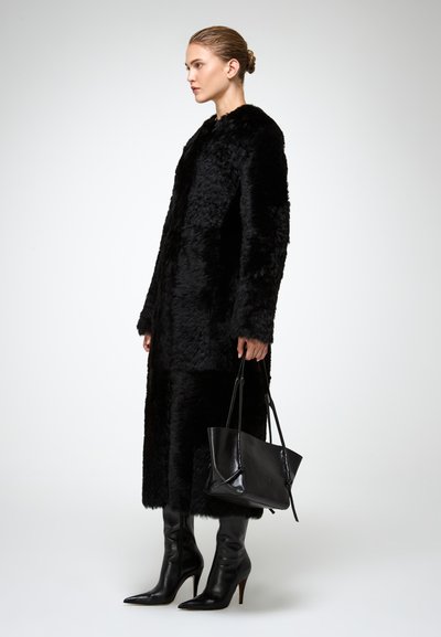 Cappotto in pelliccia sintetica nera con maniche lunghe, lunghezza midi, abbinato a stivali al ginocchio in pelle nera aderenti e una borsa strutturata nera.