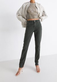 Jeans skinny verde oliva con un diseño clásico de cinco bolsillos, combinados con una chaqueta bomber beige corta y una blusa de textura clara.