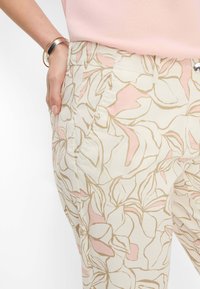 Pantalons légers avec une base beige clair, présentant un motif floral en rose doux et or. Des poches latérales et une coupe décontractée sont visibles.