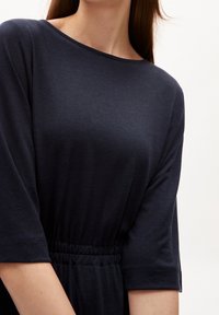 Tummansininen mekko, jossa on kyynärpäihin ulottuvat hihat, pyöreä pääntie ja vyötärölle kerätty yksityiskohta. Materiaali näyttää sileältä, pehmeältä ja hieman laskeutuvammalta.