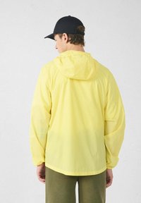 Lichtgele regenjas met capuchon, gemaakt van een glad, lichtgewicht materiaal. Voorzien van elastische manchetten en een ontspannen pasvorm.