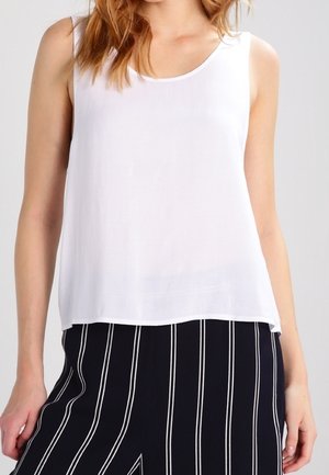 Femme portant un haut blanc sans manches et un pantalon noir avec des rayures blanches verticales, montrée du cou jusqu'au milieu des cuisses sur un fond uni.