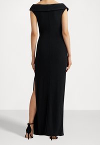 Schwarzes Maxikleid mit schulterfreiem Design, taillierter Passform, seitlichem Schlitz und glatter Textur. Kombiniert mit schwarzen High-Heels.