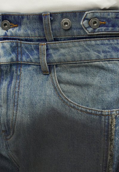 Κοντινό πλάνο στη μέση ενός μπλε τζιν παντελονιού που δείχνει τρία μεταλλικά κουμπιά και θηλιές ζώνης με ορατές ραφές.