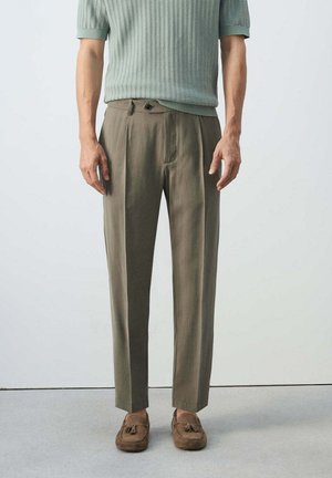 Mann trägt hellgrünes kurzärmliges Strickoberteil, olivfarbene plissierte Hose und braune Wildleder-Tassel-Loafers, steht vor einfachem Hintergrund.