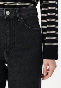 Black denim high-waisted jeans with a front button closure, five-pocket design, and a subtle textured finish. Striped long-sleeve shirt visible.