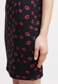 Jupe noire ajustée en tissu texturé, avec un motif de marques de lèvres rouges réparties uniformément sur la surface. Finition lisse, longueur genoux.