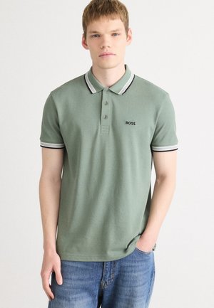 Ung mand iført en lysegrøn polo-shirt med sort og hvid kant samt BOSS-logo, iført blå jeans, stående mod en ensfarvet baggrund.