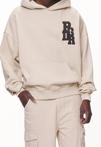 Beige huva sweatshirt gjord av mjukt tyg, med en kenguruficka och en staplad broderad logga i mörkbrunt på bröstet.