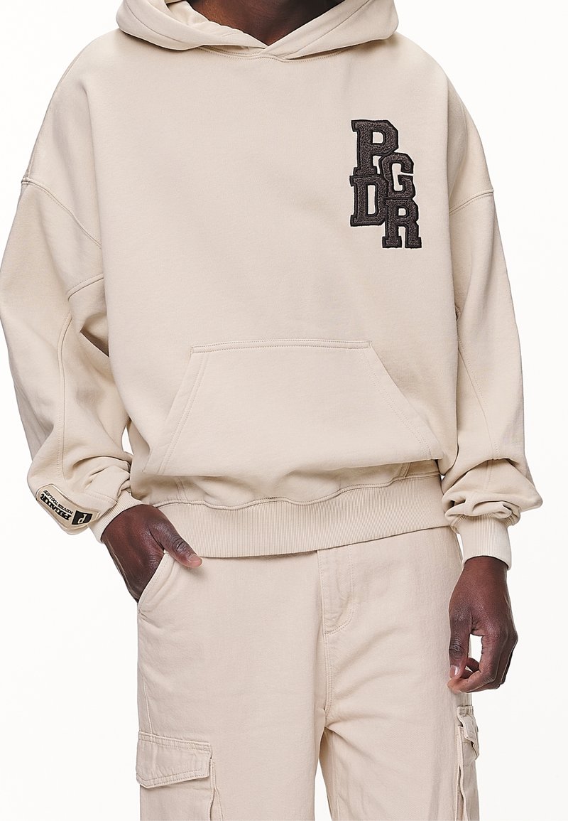 Beige huva sweatshirt gjord av mjukt tyg, med en kenguruficka och en staplad broderad logga i mörkbrunt på bröstet.