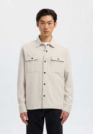 Hombre con camisa beige clara abotonada con dos bolsillos de solapa en el pecho y pantalones negros sobre un fondo blanco liso.