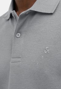 Grå polo t-shirt lavet af tekstureret stof, med kravehalsudskæring, to knapper og et diskret logo på brystet.