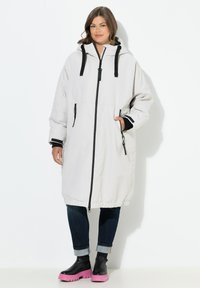 Lange lichtgrijze parka met zwarte accenten, ritsluiting, trekkoordkap, zijzakken en elastische manchetten, gedragen over donkere jeans en zwarte laarzen met roze zolen.