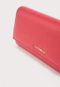 Rood textuurleer portemonnee met gouden "Coccinelle" logo aan de voorkant, weergegeven vanuit een hoek op een plain witte achtergrond.