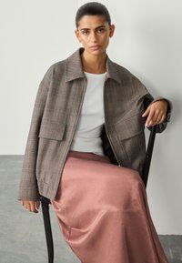 Giacca oversize marrone a quadri con grandi tasche, indossata sopra un top a coste bianco, abbinata a una maxi gonna in satin rosa. Tessuti morbidi e design a strati.