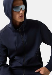 Navy modrá zipová mikina s vysokým límcem, dvěma bočními kapsami a stahovacími šňůrkami. Model má na sobě reflektivní sluneční brýle. Hladká, měkká látka.