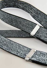 Svarta och silverfärgade vävda elastiska hängslen med metallklämmor på en vit yta, arrangerade i loopade och överlappande linjer.