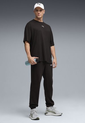 Maglietta nera oversize con logo bianco, pantaloni della tuta neri e scarpe da ginnastica grigie/verdi. Tiene in mano una bottiglia d'acqua azzurro chiaro. Cappellino bianco.