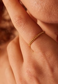 Bague en or avec un design perlé, portée sur un doigt. La surface est lisse et brillante contre la peau texturée.