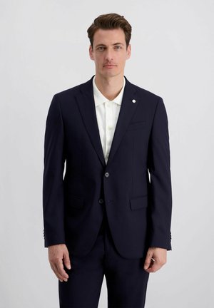 Man met bruin haar, gekleed in een donkerblauw colbert over een wit overhemd met knopen, staand tegen een effen lichtgrijze achtergrond.