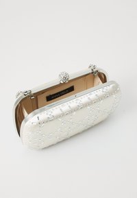 Bijela clutch torba s teksturiranim vanjskim dijelom, srebrnim akcentima i draguljastim kopčanjem. Unutrašnjost je obložena mekim materijalom. Vidljiva je potpisna oznaka.