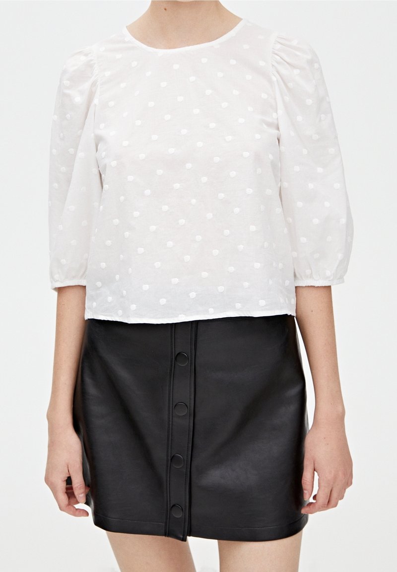 Femme portant un chemisier blanc à texture à pois avec des manches bouffantes trois-quarts et une mini-jupe noire en cuir avec boutons sur le devant.