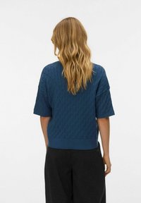 Plavi pleteni džemper s teksturiranim uzorkom, kratkim rukavima i rebrastim rubom. Nošen s tamnim, širokim hlačama.