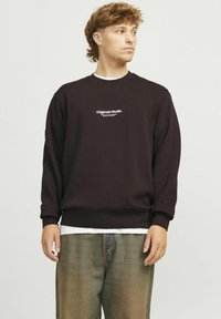 Donkerbruin sweater met een ronde hals, met witte tekst "Originals Studio" op de borst. Soepele textuur, ribgebreide boorden en zoom.
