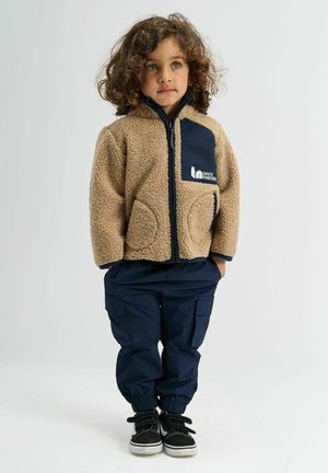 Bežová fleecová bunda s námorníckymi akcentmi, predným zipsom a vreckami na ruky, skombinovaná s námorníckymi cargo nohavicami a čiernymi teniskami s šnúrkami.