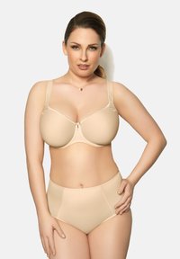 Mujer de piel clara y cabello rubio que lleva puesto un sujetador con aros beige y bragas de cintura alta, posando contra un fondo liso claro.